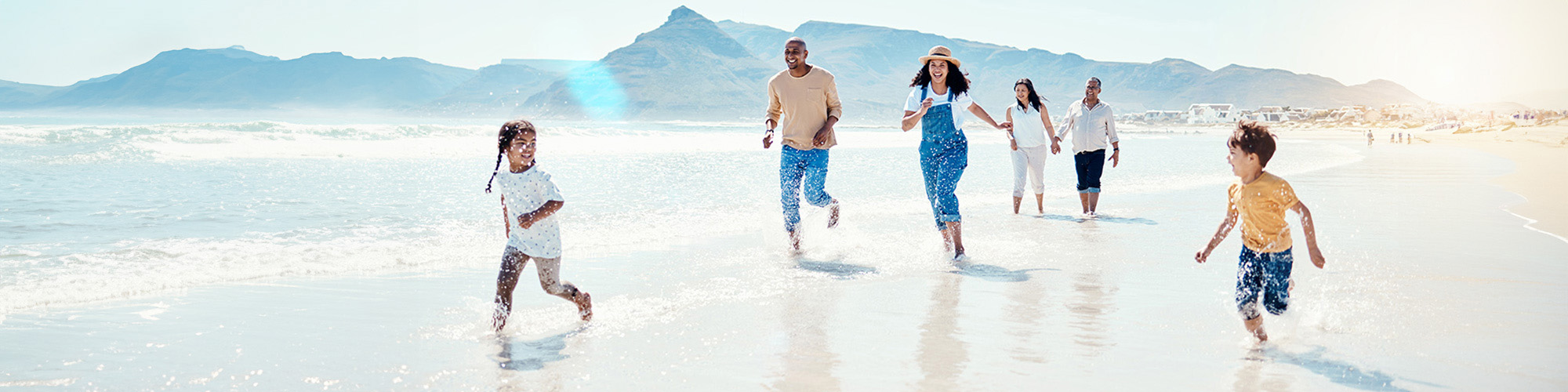 Eine Familie genießt gemeinsam den Tag am Strand. Zwei Kinder rennen lachend durchs seichte Wasser, während drei Erwachsene im Hintergrund Hand in Hand und lächelnd am Ufer entlanglaufen. Im Hintergrund sind Berge, das Meer und einige Häuser zu sehen, die Sonne scheint hell.