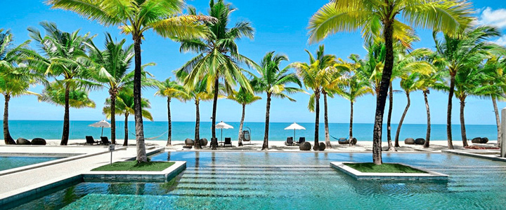 Kristallklarer Pool, umgeben von zahlreichen Palmen im Sonnenlicht, Blick auf Strand und Meer.