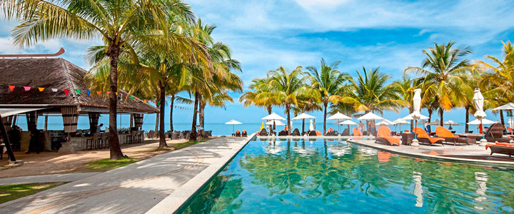 Türkisfarbener Pool, umgeben von zahlreichen Palmen im Sonnenlicht, Blick auf Strand und Meer.