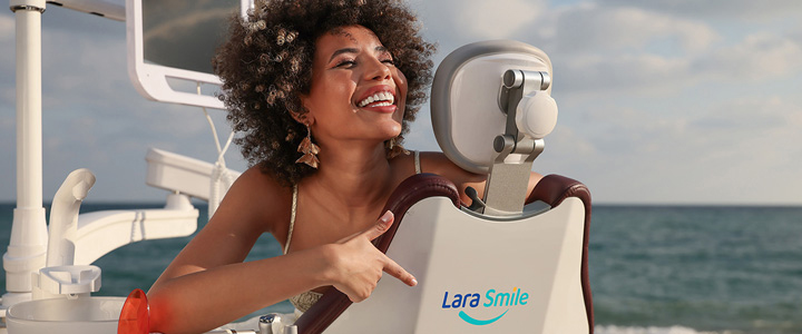 Frau sitzt im Zahnarzt-Behandlungsstuhl am Strand und lächelt, während sie auf das „Lara Smile“-Logo zeigt.