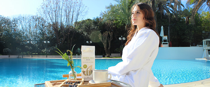 Frau steht in einem Bademantel an einem Tisch vor einem Pool mit kristallklarem Wasser und blickt nach Links in Richtung der Sonne.