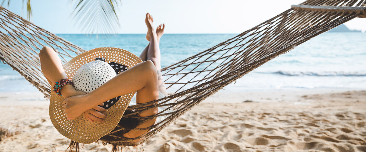 Junge Frau startet ganz entspannt in einer Hängematte am Strand in ihren Sommerurlaub - Die Reise ganz nach ihren Wünschen und ohne Stress geplant.