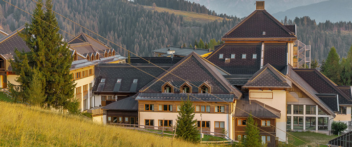 Das Aldiana Club Schlanitzen Alm zeigt eine Außenansicht mit viel Holz und natürlicher Atmosphäre. Davor erstreckt sich eine sommerliche Wiese, im Hintergrund liegen bewaldete Bergzüge.