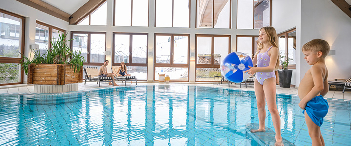 Großzügiger Innenpool, der sich über die gesamte Bildbreite erstreckt, in einer offenen, lichtdurchfluteten Raumgestaltung. Im Hintergrund entspannte Eltern auf Liegestühlen, während die Kinder bereit zum Sprung ins Wasser stehen.