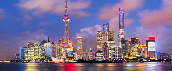 Leuchtende Skyline von Shanghai bei Nacht mit spiegelnden Lichtern auf dem Wasser davor und tief dunkelblauem Himmel im Hintergrund.