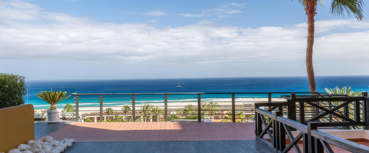 Moderne Außenterrasse mit dunklem Bodenbelag und Glasreling, die einen ungehinderten Panoramablick auf den hellen Sandstrand und das türkisfarbene Meer eröffnet.