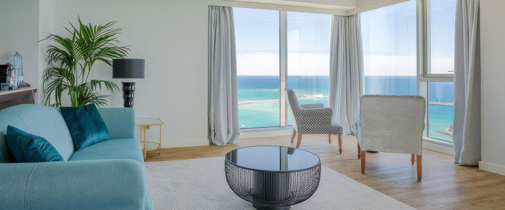 Modernes, helles Wohnzimmer mit einem hellblauen Sofa, einem runden Designer-Couchtisch und großen Panoramafenstern, die einen weiten Blick auf das türkisfarbene Meer bieten.