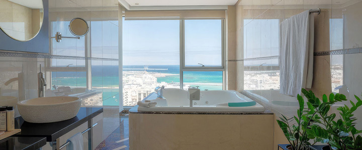 Luxuriöses Badezimmer mit einer großen Whirlpool-Badewanne direkt am Fenster, Marmorboden und einem modernen Aufsatzwaschbecken, mit Blick auf die Küste und das Meer.