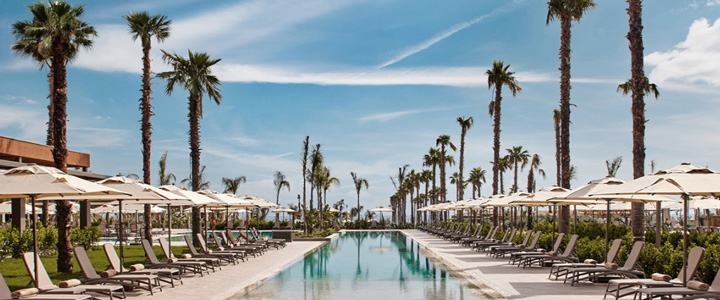 Im Zentrum liegt ein türkis schimmernder Pool, umgeben von Sonnenliegen und hohen Palmen. Unter klarem, blauem Himmel fällt helles Sonnenlicht auf die gepflegte Anlage.