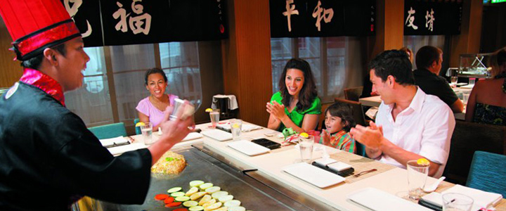 Eine Familie sitzt am Teppanyaki-Grill der NCL Epic, während der Koch die Speisen auf der glänzenden Edelstahlplatte zubereitet.