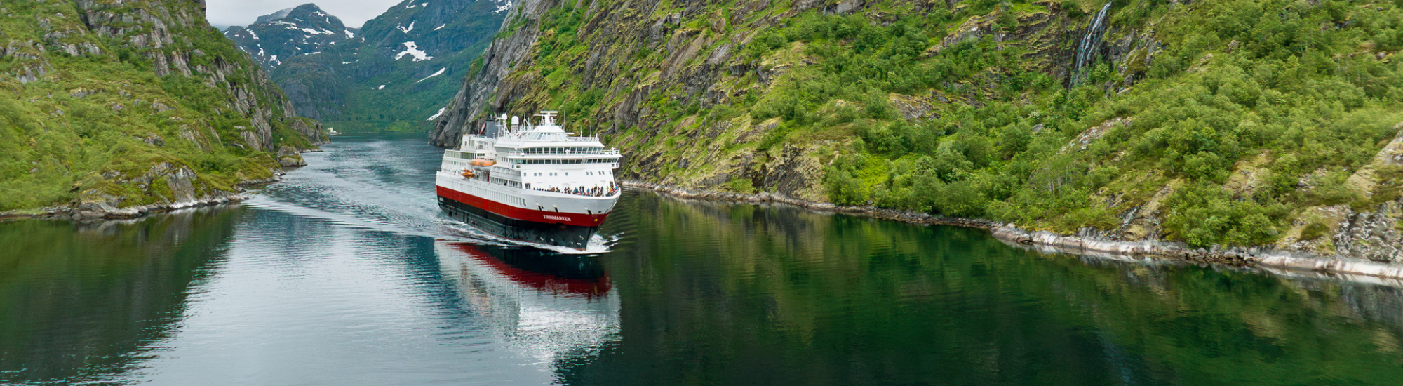 Hurtigruten Landingpage Trollfjorden_Drone_HGR_168148_Photo_Espen_Mills (2000 x 550 px) Die nickoSPIRIT gleitet im warmen Abendlicht der untergehenden Sonne über den Fluss, während das Wasser goldene Reflexe trägt, Time to discover.